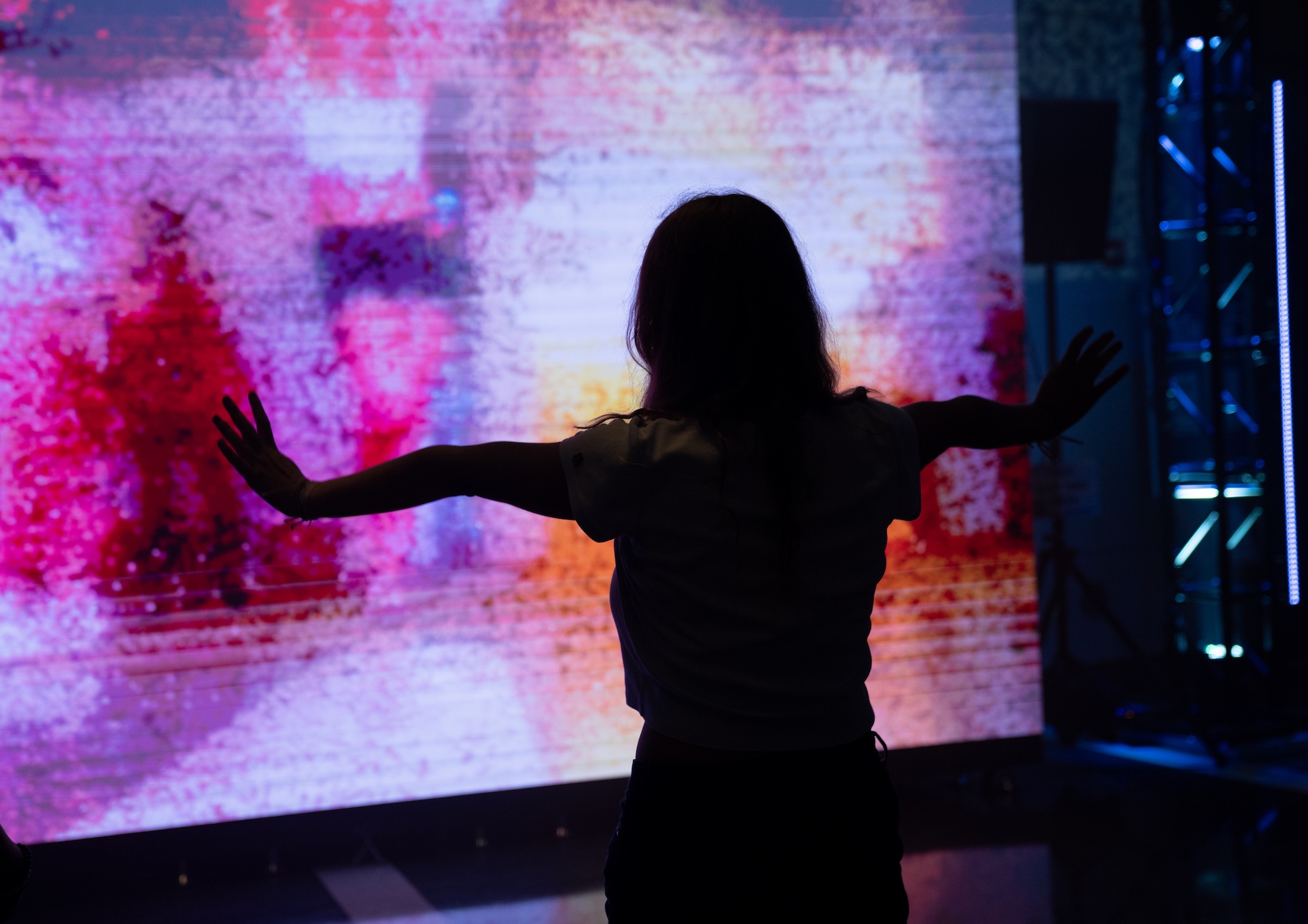 A silhouetted figure stands with arms outstretched before a large-scale interactive projection display at NextNOW Fest, engaging with vibrant pink, purple, and orange abstract visuals that respond to movement. Blue LED panels are visible on the right side of the darkened space. The immersive installation creates a dynamic fusion of human gesture and digital art in the Grand Pavilion.