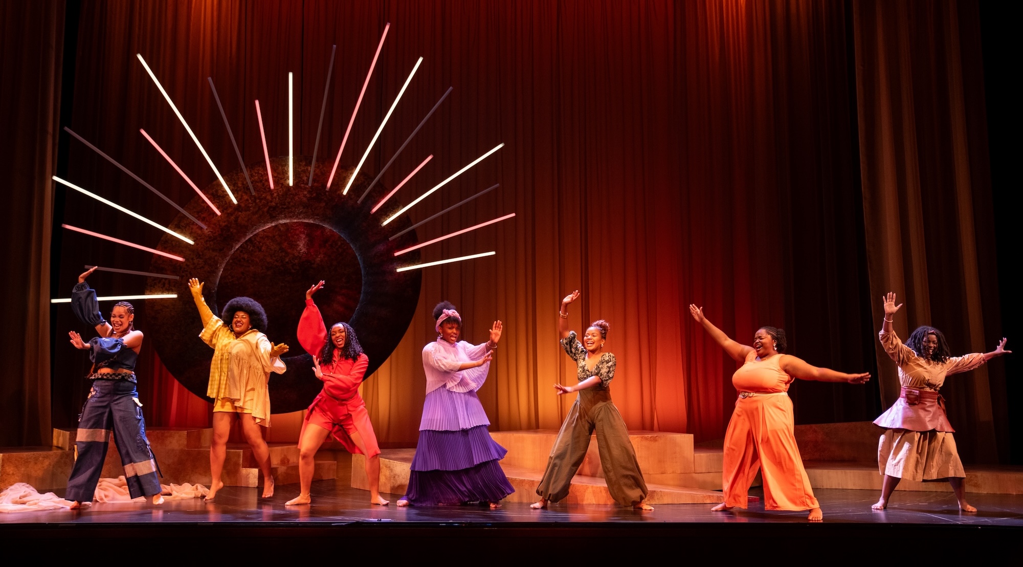 Seven performers in vibrant costumes strike joyful, expressive poses across the stage in a production of "for colored girls," with arms outstretched in celebration against rich red curtains. A striking sunburst lighting sculpture radiates behind them as the dramatic focal point. The cast wears an array of colorful outfits including yellow, red, purple, and earth tones, capturing the show's powerful emotional journey.